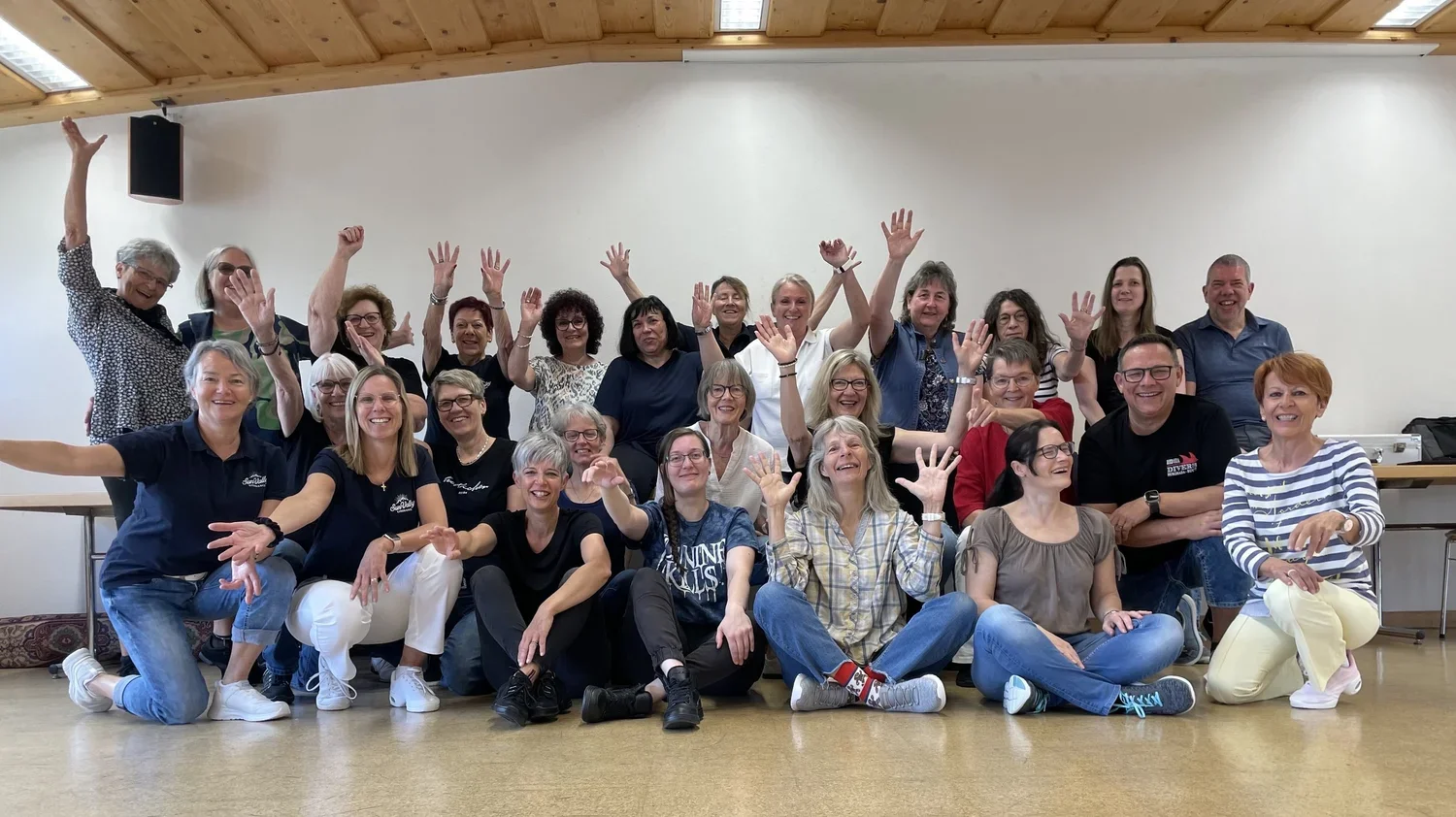 Gruppe von lachenden Menschen in einem Raum, einige sitzen auf dem Boden, andere stehen im Hintergrund, alle haben die Hände hoch in die Luft gestreckt.