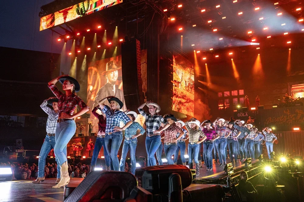 Line dancing Show mit Frauen in Cowboyhüten und Karomustern auf der Bühne bei einem Konzert, mit bunten Lichtern und großen Leinwänden im Hintergrund.