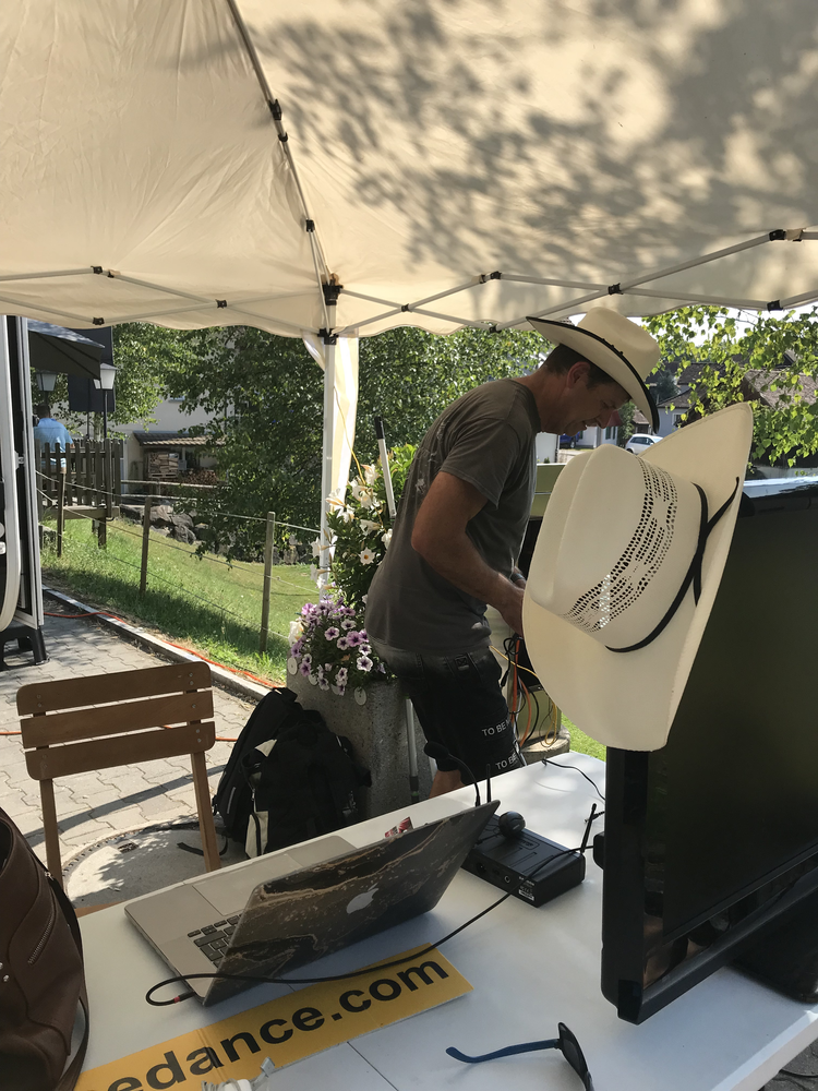 Ein Mann steht unter einem Sonnenschirm im Freien, arbeitet an technischen Geräten. Der Tisch hat einen Laptop, Bildschirm und einen gelben Aufkleber mit der Webseite ‘dance.com’. Es gibt auch Hüte auf einem Ständer und eine Tafel mit Blumen im Hinte