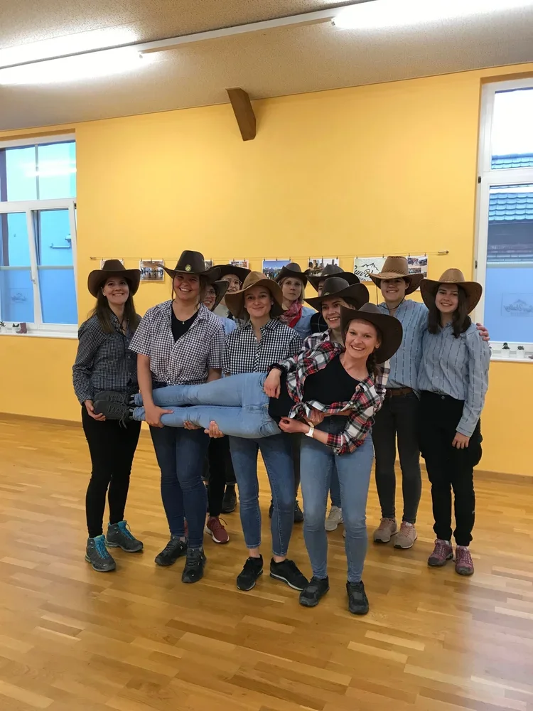 Eine Gruppe von jungen Frauen trägt Cowgirl-Hüte und Westernkleidung, während sie eine Person horizontal in der Mitte halten, in einem Raum mit gelben Wänden und Holzfußboden.