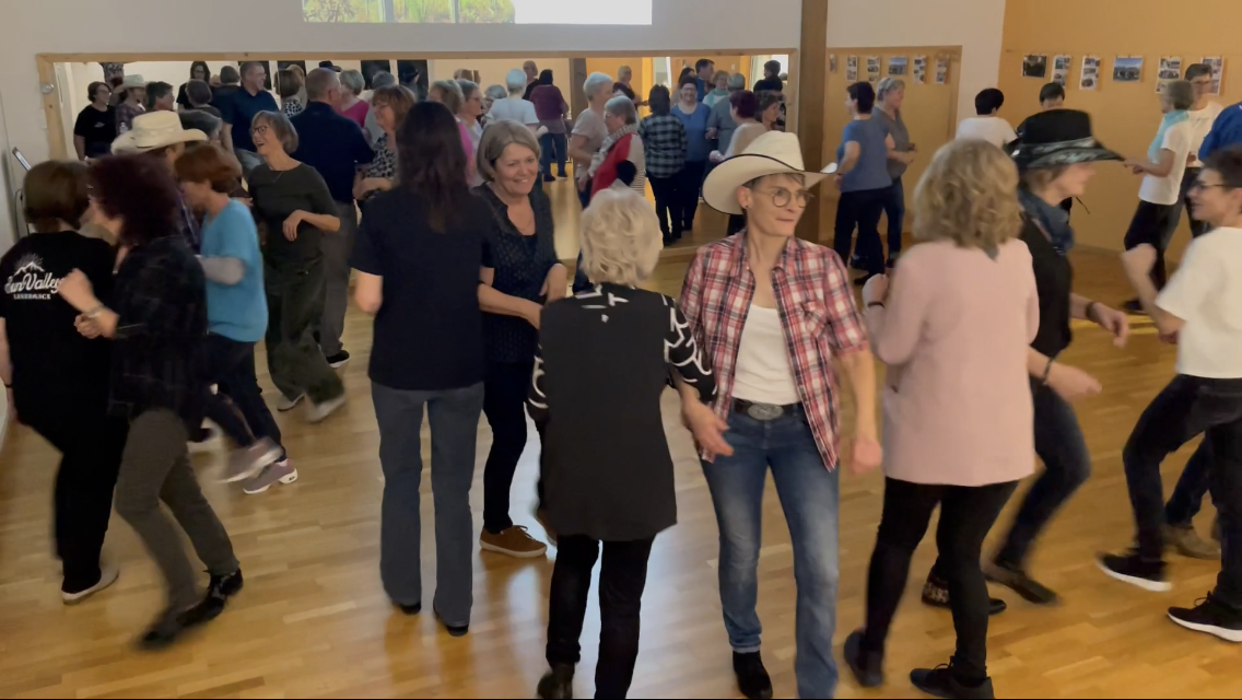 Menschen tanzen in einer Gemeinschaftshalle, viele lachen und haben Spaß, mit Holzwänden und einer Fotowand im Hintergrund.