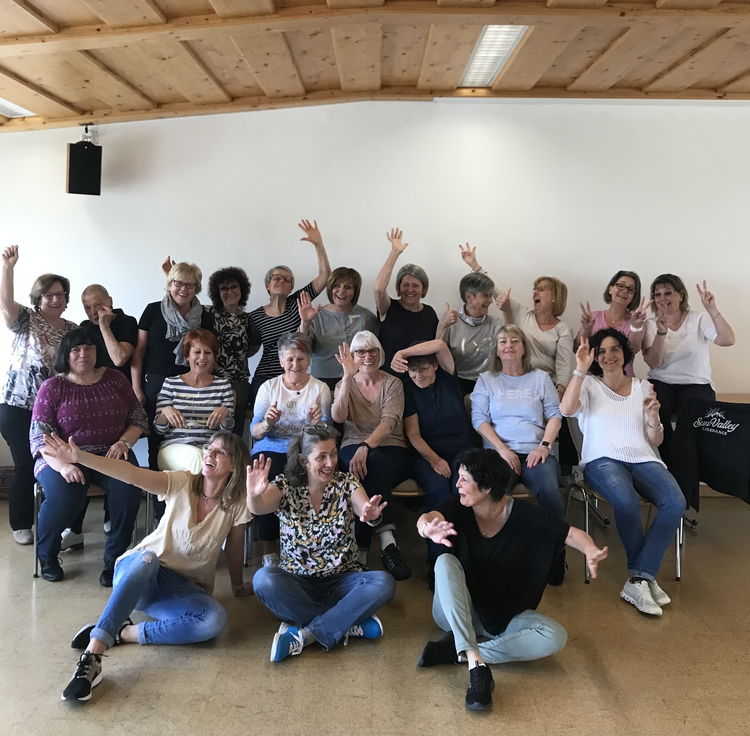 Eine Gruppe von Frauen lächelt und winkt in einem Raum mit Holzdecke und weißer Wand