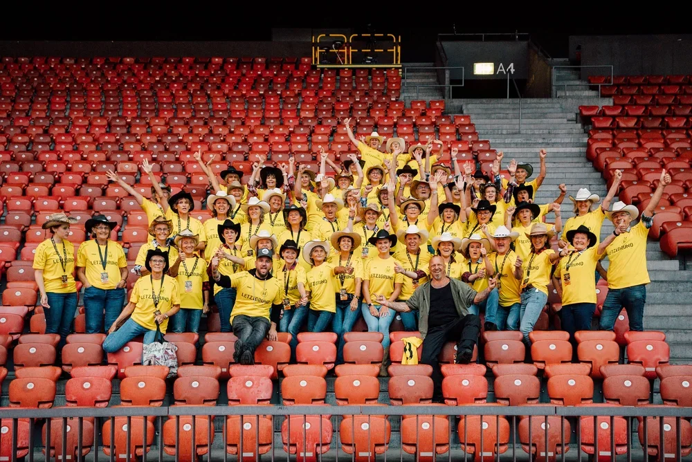 Gruppe von Menschen in gelben T-Shirts und Hüten, die in einem leeren Stadion auf den roten Sitzen posieren und jubeln.