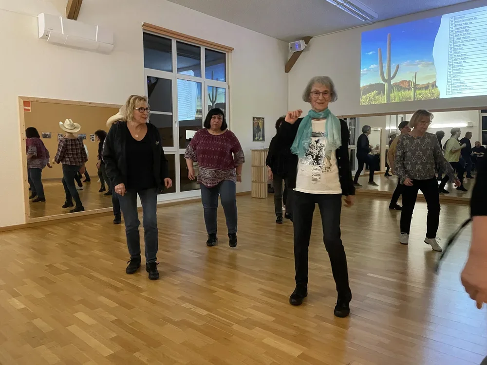 Eine Gruppe von Menschen tanzt in einem Saal, der Holzboden hat und eine große Spiegelwand hat, im Hintergrund ist eine große Projektion mit Cactus auf der Wand.