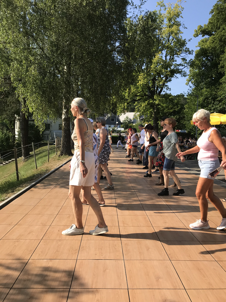 Eine Gruppe älterer Menschen tanzt im Freien an einem sonnigen Tag auf einer Holzterrasse, umgeben von grünen Bäumen.