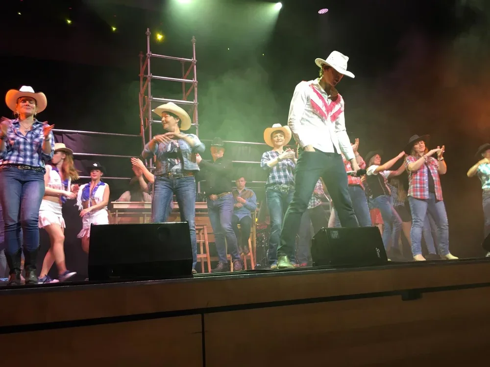 Gruppe von Darstellern in Cowboystil auf einer Bühne bei einer Theater- oder Tanzaufführung, mit Scheinwerferbeleuchtung und Kulisse im Hintergrund.