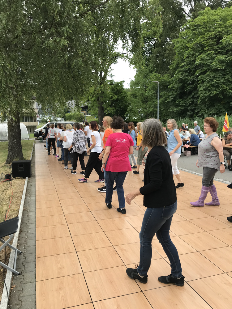 Menschen tanzen im Freien auf einer Holzterrasse bei einem Gemeinschaftsfest, umgeben von Bäumen.