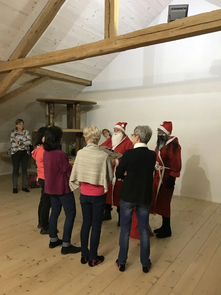 Menschen in einem Raum mit Holzdekor, einige verkleidet als Weihnachtsmänner, andere bekannte Personen. Es scheint eine festliche Weihnachtsveranstaltung zu sein.