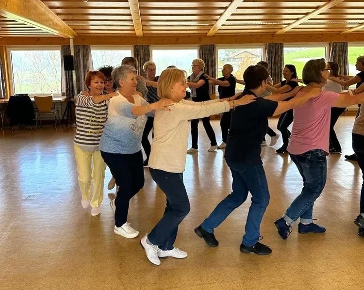 Ältere Menschen tanzen in einer Gruppe, die Arme auf den Schultern der Person vor ihnen. Das Treffen findet in einem Gemeinschaftsraum mit Holzbalken und großen Fenstern statt.