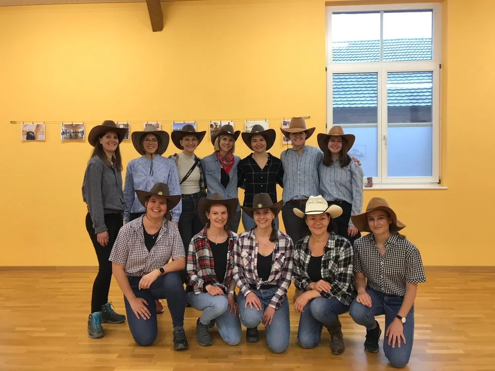 Gruppe von zehn Frauen in Cowboyhüten und Westernkleidung in einem Raum mit gelber Wand und Fensterscheibe.