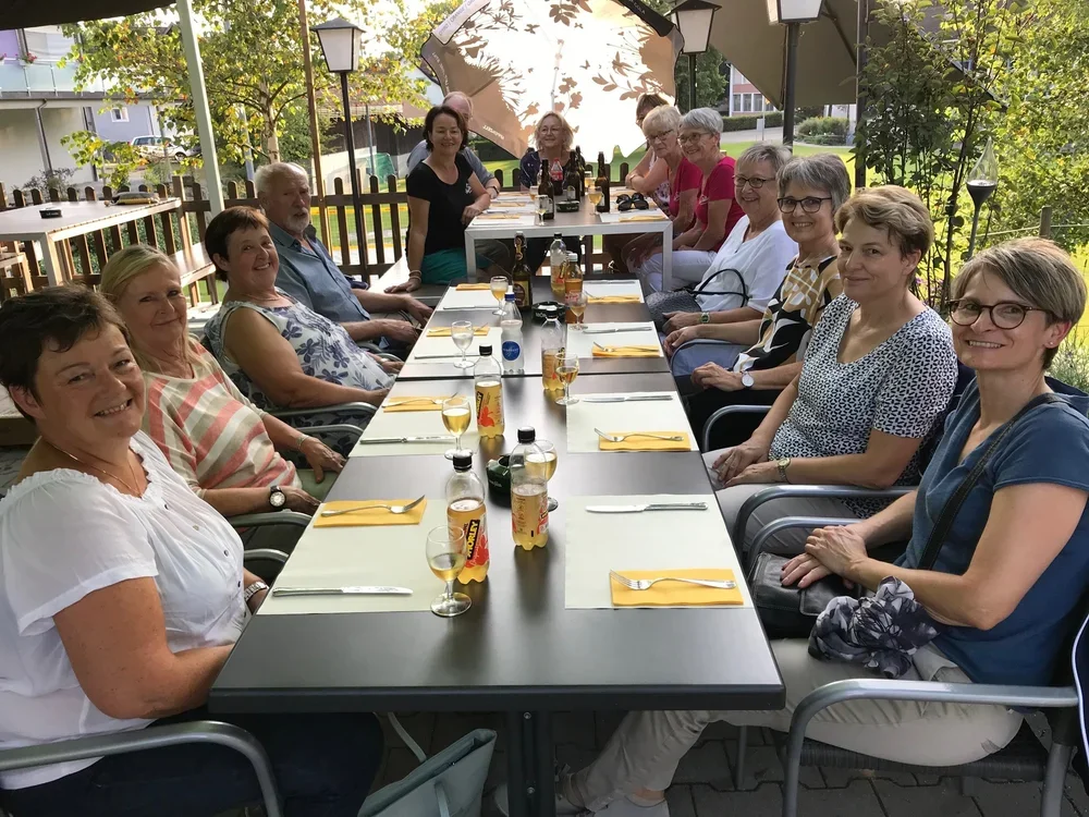 Eine Gruppe älterer Menschen sitzt an einem langen Tisch im Freien bei einem Treffen oder Essen.