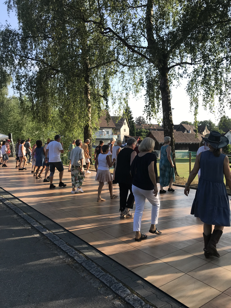 Menschen, die im Freien auf einer Tanzfläche tanzen, mit Bäumen und Häuser im Hintergrund an einem sonnigen Tag.