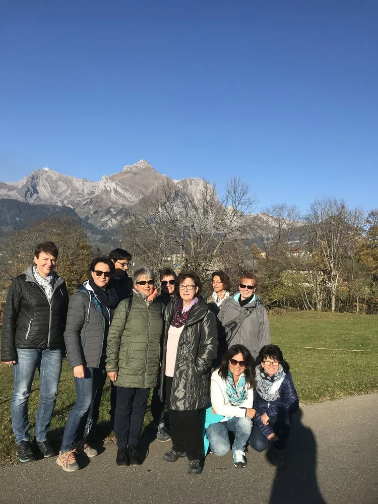 Eine Gruppe von zehn Menschen steht vor einer Berglandschaft mit Bergen im Hintergrund und blauen Himmel, vermutlich bei sonnigem Wetter.