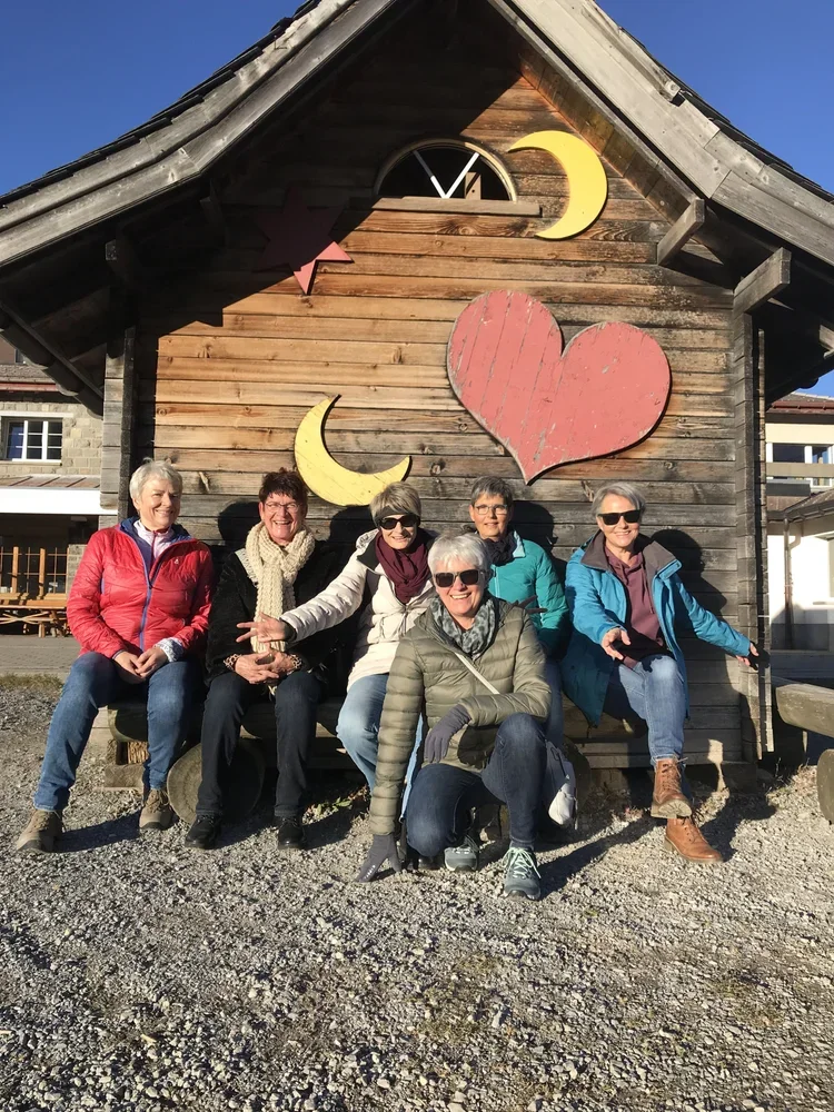 Sechs Frauen sitzen vor einer Holzhütte mit Sonnen- und Monden-Dekorationen an der Wand, sie lachen und genießen die Sonne, bei gutem Wetter.