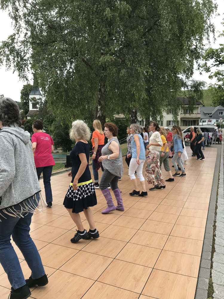 Menschen tanzen auf einer Holzterrasse im Freien, im Hintergrund ein großer Baum und einige Häuser.