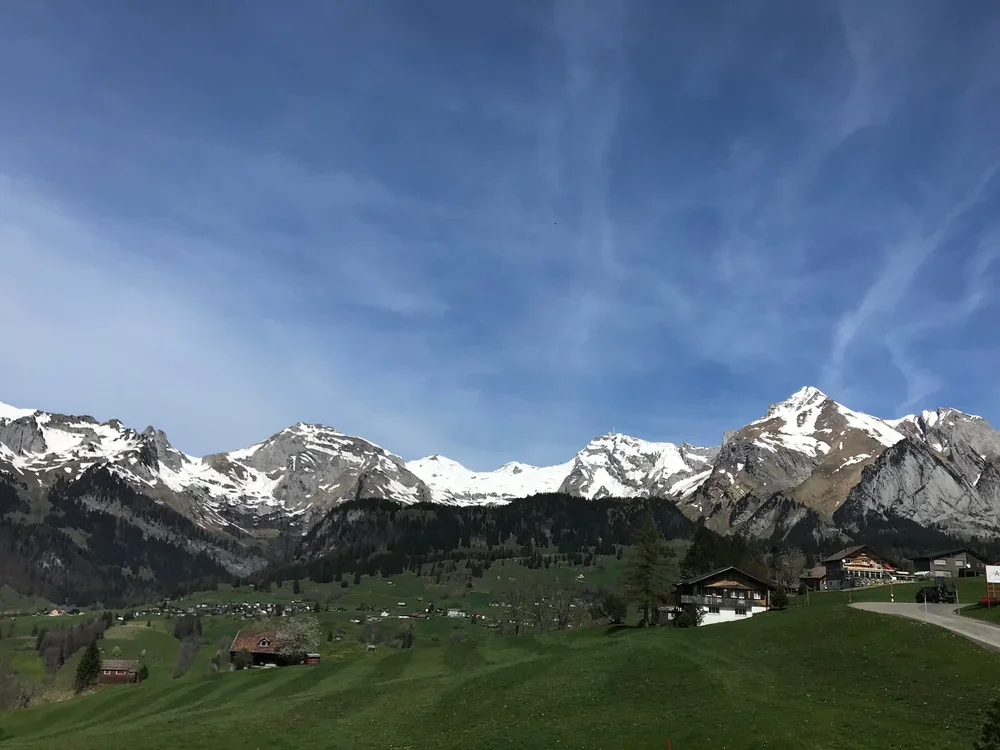 Berglandschaft mit schneebedeckten Gipfeln, grünen Wiesen und einigen Häusern im Tal, unter einem blauen Himmel mit Wolken.