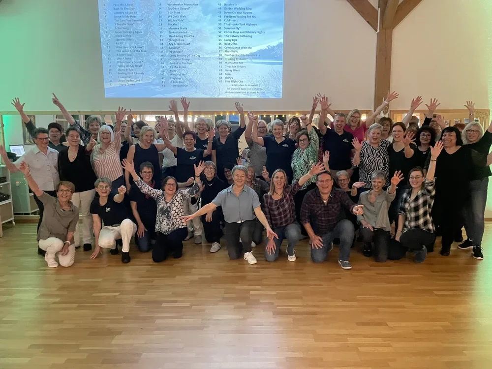 Eine große Gruppe lachender Menschen, die in einem Raum posieren, mit erhobenen Händen, vor einer großen Leinwand mit einem Song-Playlist. Der Raum hat Holzboden und ist hell erleuchtet.