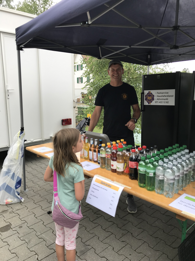 Ein Junge verkauft Getränke an einem Stand mit Kindern, unter einem blauen Pavillon, im Freien, mit Gebäuden und Bäumen im Hintergrund.