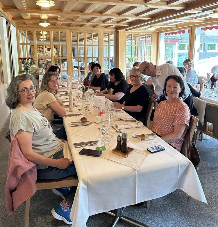 Menschen sitzen bei einem gemütlichen Essen an einem langen, elegant gedeckten Tisch in einem lichtdurchfluteten Raum mit Holzbalkendecke, einige lächeln in die Kamera, während andere sich unterhalten.