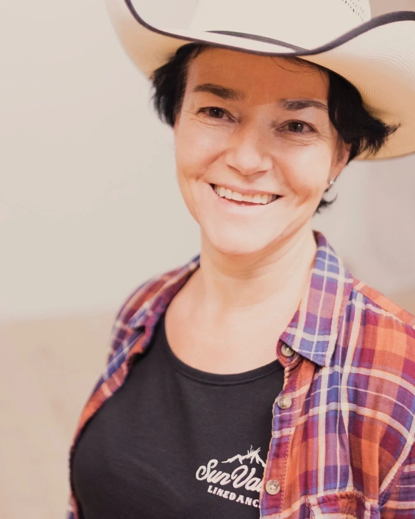 Frau mit schwarzen Haaren, trägt einen weißen Cowboyhut, ein rotes kariertes Hemd und ein schwarzes T-Shirt mit weißem Logo.