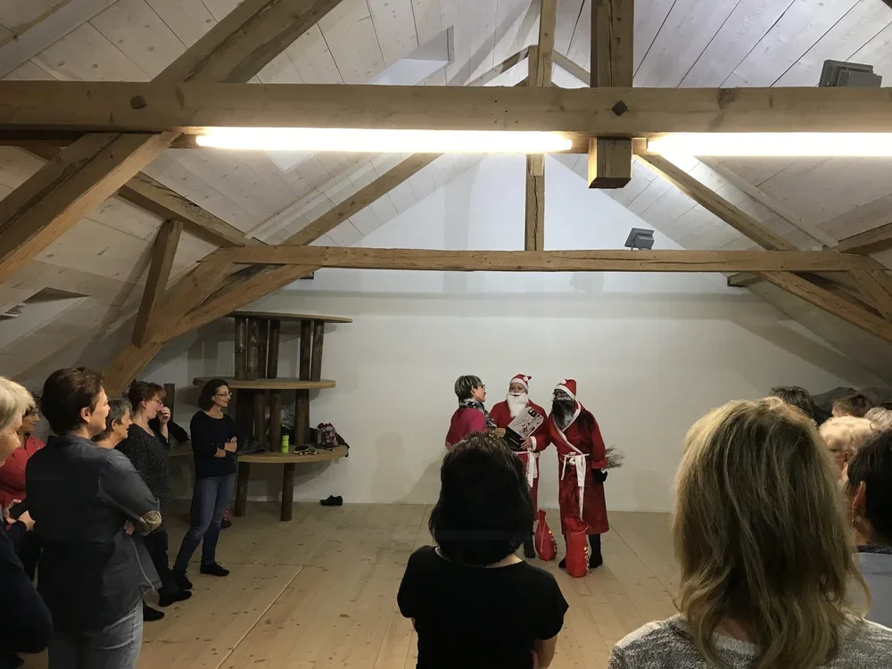 Menschen in einem Raum mit Holzdecke, unten auf der Bühne stehen zwei Personen in Weihnachtsmannkostümen, eine Person liest aus einem Buch vor, im Hintergrund sind weitere Zuschauer zu sehen.