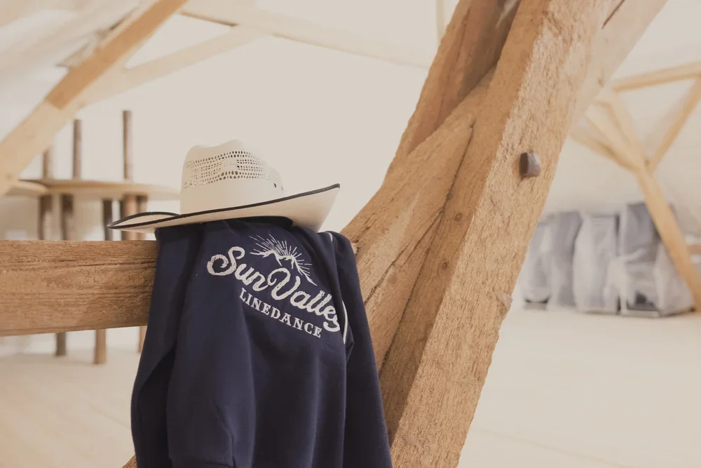 Sonnenhut und Jacke hängen an einem Holzbalken in einem Dachgeschoss, im Hintergrund einige weitere Holzbalken und Fenster.