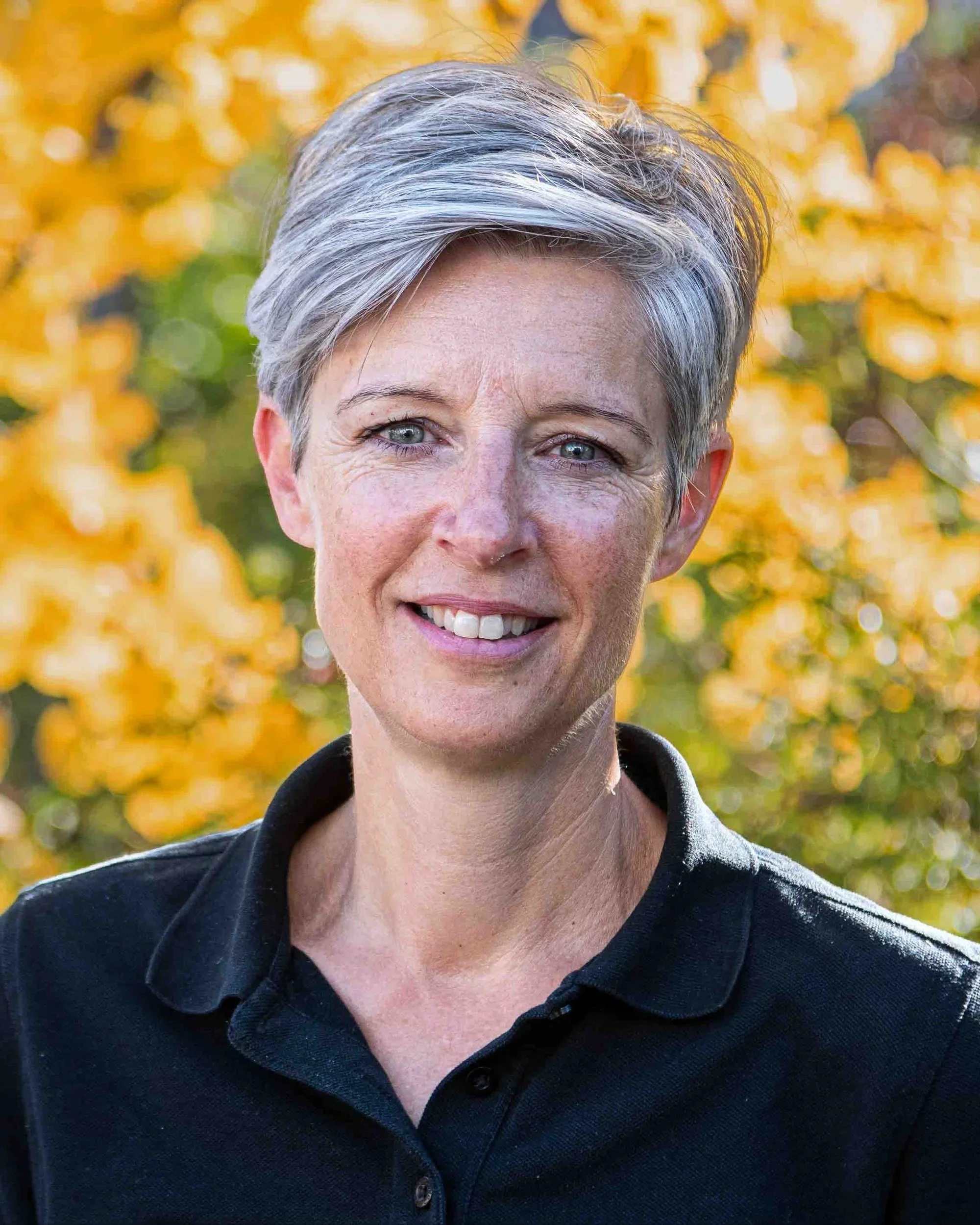 Eine Frau mit kurzen, grau-weißen Haaren und einem schwarzen Polohemd, stehend vor einem Hintergrund mit gelben Herbstblättern.