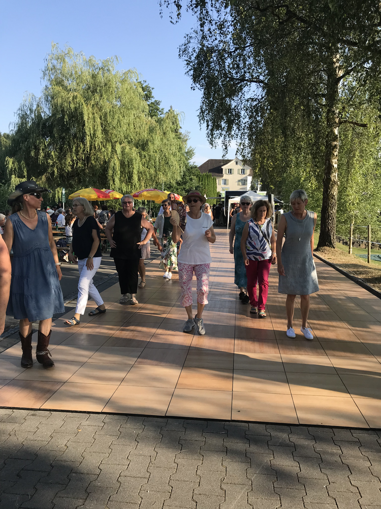 Menschen spazieren an einem sonnigen Tag durch einen Park mit Bäumen und einer Terrasse mit Sonnenschirmen.