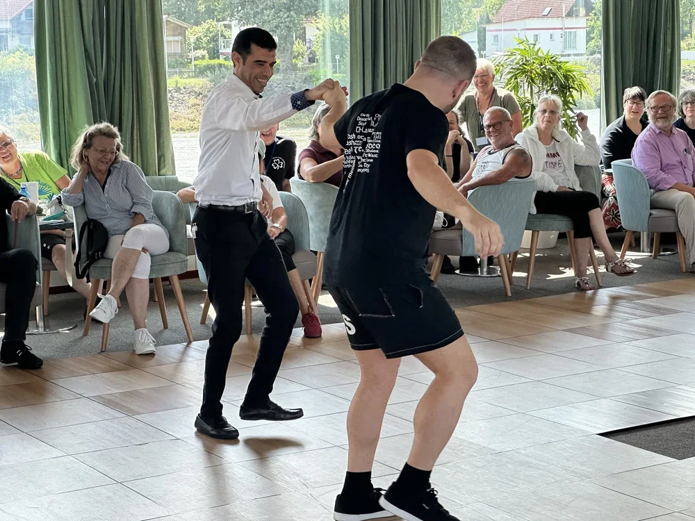 Zwei Männer tanzen auf einer Tanzfläche, während ein Publikum im Hintergrund zuschaut und lachen.
