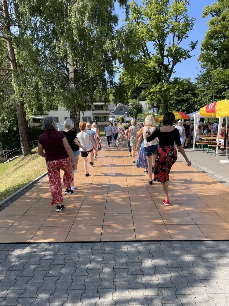 Menschen tanzen im Freien auf einem gepflasterten Weg unter Bäumen bei einer Veranstaltung mit Zelten und Ständen.