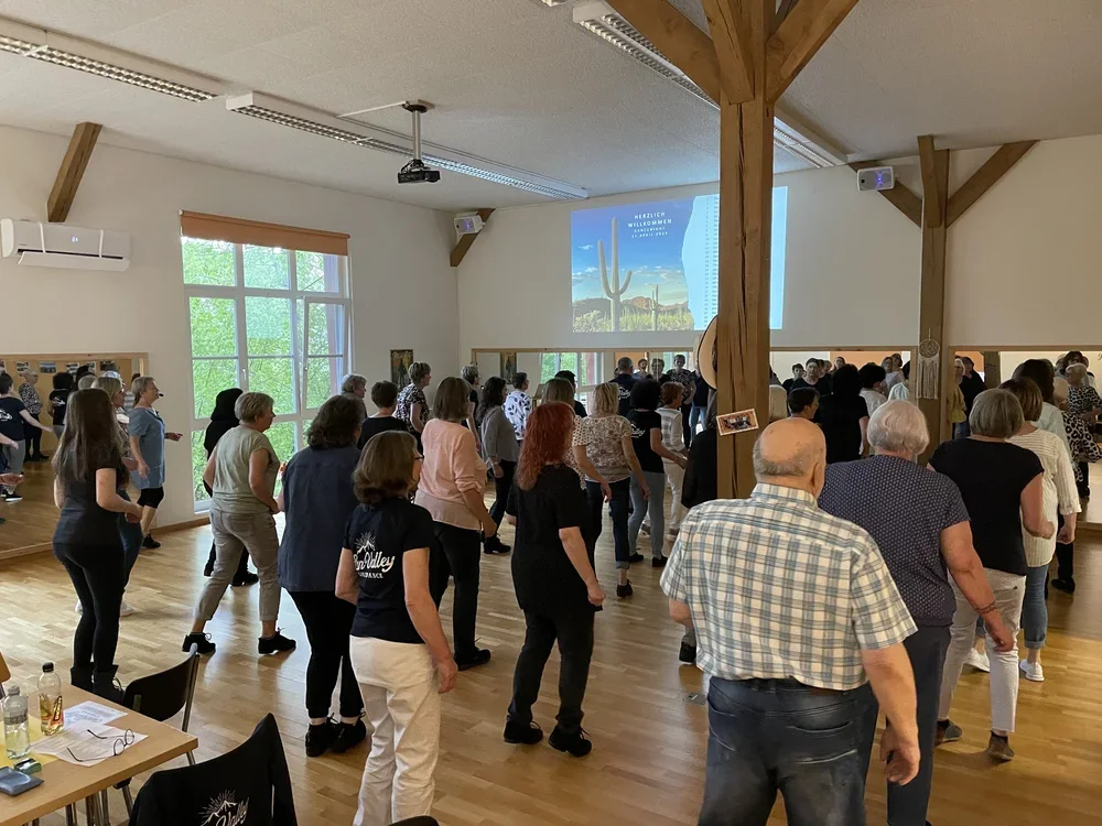 Menschen tanzen in einem hellen Raum mit großen Fenstern, einer Projektionswand mit Cactus und Wüstenlandschaft, Holzbalken und einem air conditioner.