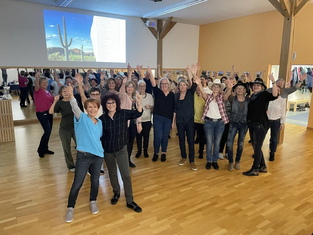 Viele lachende Frauen in einem großen Raum mit Holzfußboden, die einen GruppenFoto machen, einige haben die Arme in die Luft. Im Hintergrund ist eine große Leinwand mit einem Bild von Kakteen in der Wüste und einem Projektor sichtbar.