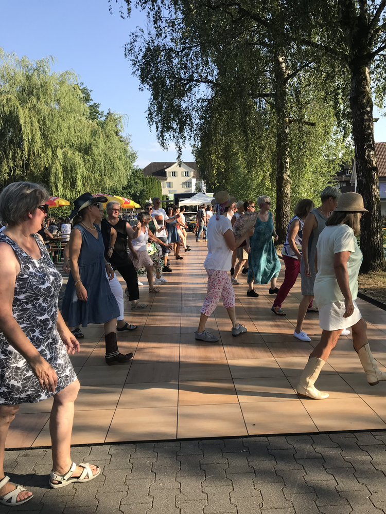 Menschen tanzen im Freien unter Bäumen an einem sonnigen Tag, einige tragen Hüte und sommerliche Kleidung.