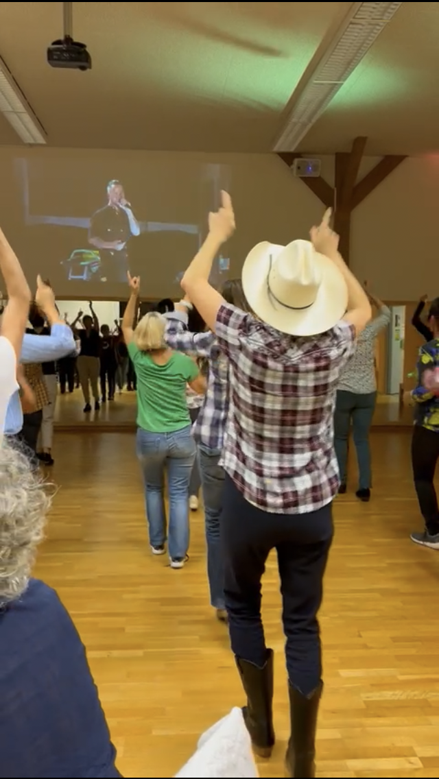Menschen in einem Saal tanzen und zur Musik auf einer großen Leinwand im Hintergrund. Ein Mann trägt einen Cowboyhut und Cowboystiefel, ein Mädchen trägt ein grünes T-Shirt, alle scheinen Spaß zu haben.