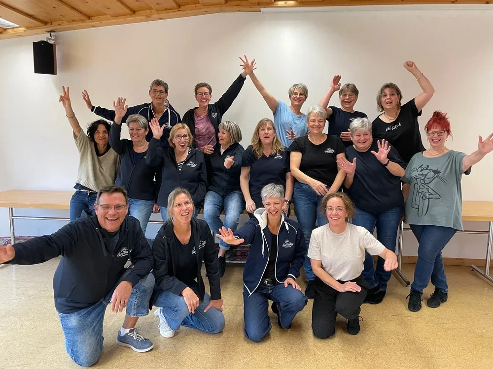 Eine Gruppe von 14 lachenden Menschen, meisten Frauen, posieren fröhlich im Raum, einige mit erhobenen Händen und Armen.