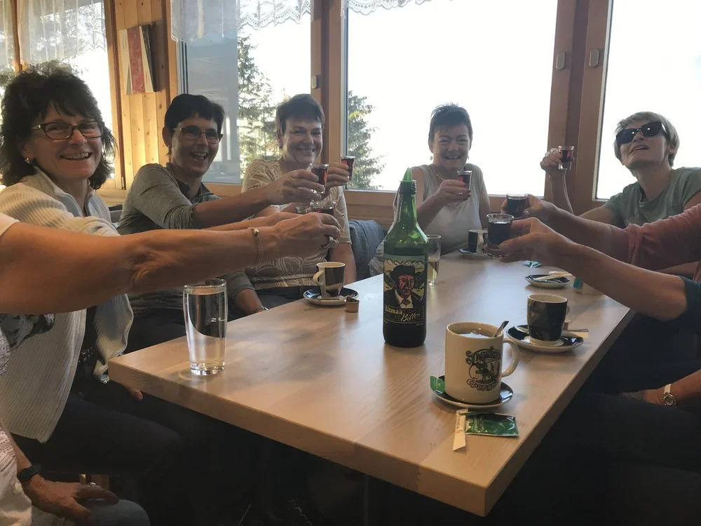 Eine Gruppe von sieben Frauen sitzt um einen Holztisch in einem Raum mit großen Fenstern. Sie heben ihre Gläser mit dunklen Getränken zum Anstoßen und lachen. Auf dem Tisch stehen Tassen, ein Wasser Glas, eine Flasche mit einem bunten Etikett, und kl