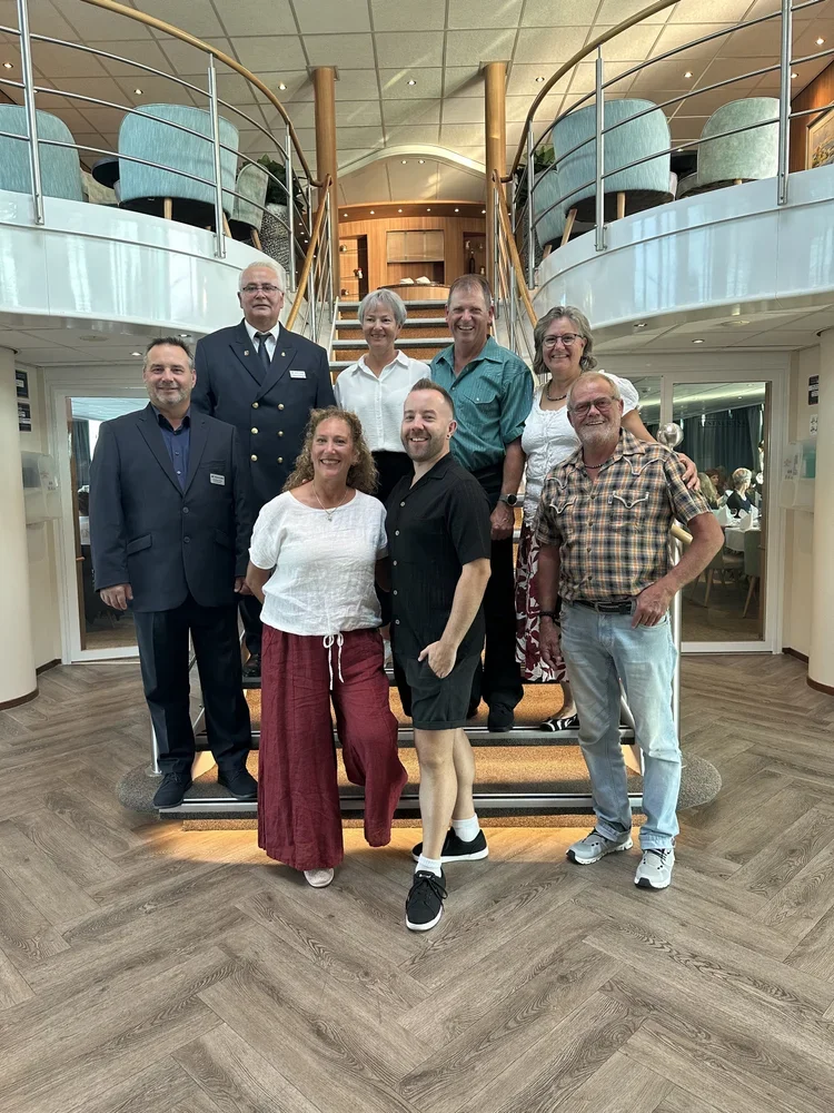 Eine Gruppe von neunzehn Personen posiert auf einer Treppe in einem gehobenen Restaurant oder Hotel. Es ist eine Mischung aus Männern und Frauen, einige in formelle Kleidung, andere in legere Kleidung. Die Umgebung ist stilvoll mit Holzdetail, modern