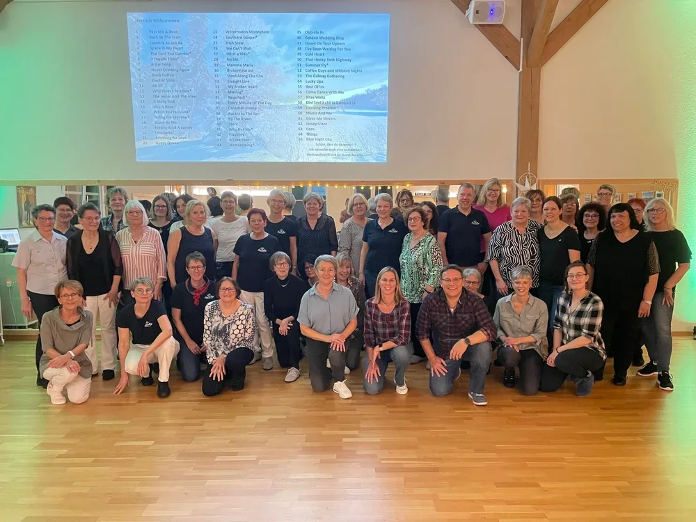 Gruppe von ca. 34 Menschen, die in einem Veranstaltungsraum vor einer großen Leinwand stehen. Die Leinwand zeigt eine Playlist oder Präsentationsliste.