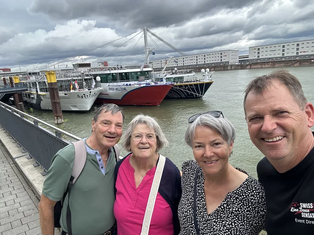 Vier Personen posieren für ein Gruppenfoto am Hafen, mit mehreren Schiffen und modernen Gebäuden im Hintergrund, bei dunklem Himmel.