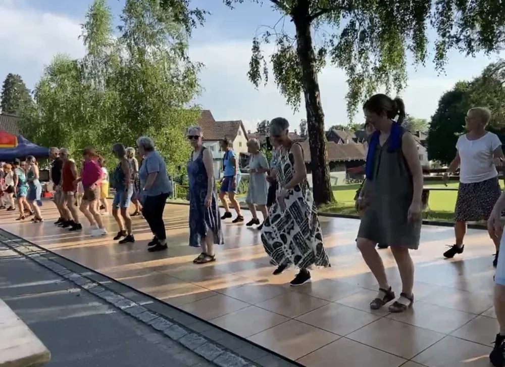 Menschen tanzen im Freien an einem sonnigen Tag auf einer Holzterrasse, umgeben von Bäumen und Häusern im Hintergrund.