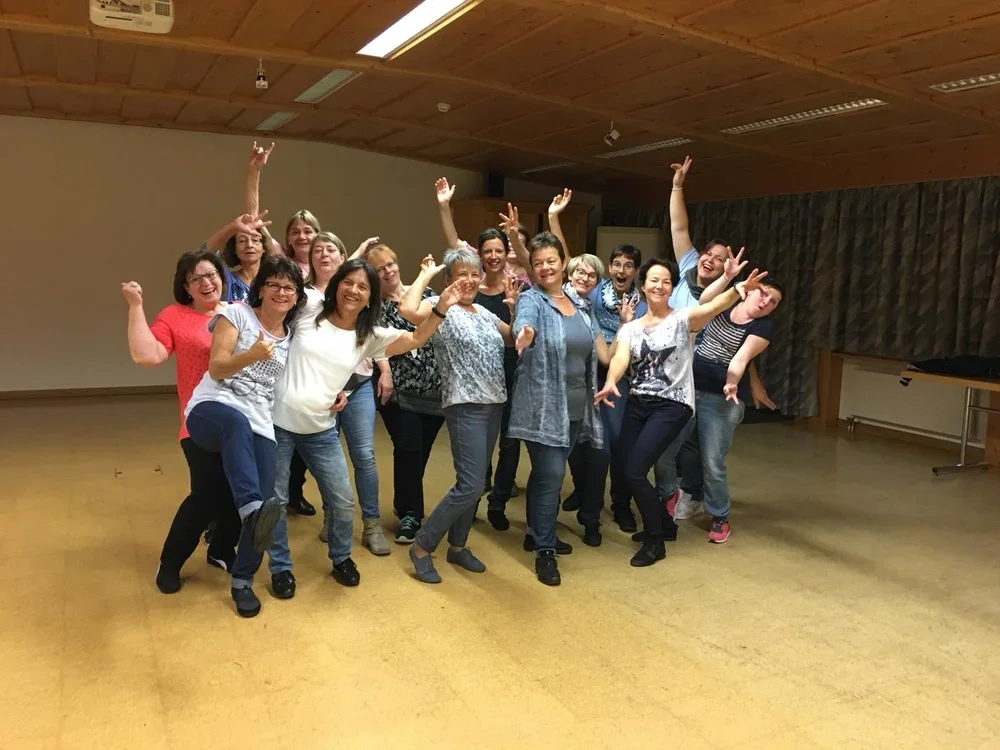 Gruppe lachender Frauen und Männer in fröhlicher Pose in einem Raum mit Holzdecke.