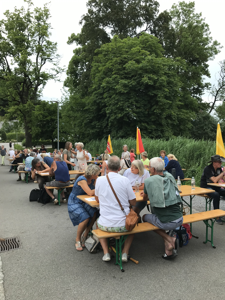 Menschen sitzen an Tischen im Freien bei einer Veranstaltung im Park, einige unter roten und gelben Schirmen, umgeben von grünen Bäumen.
