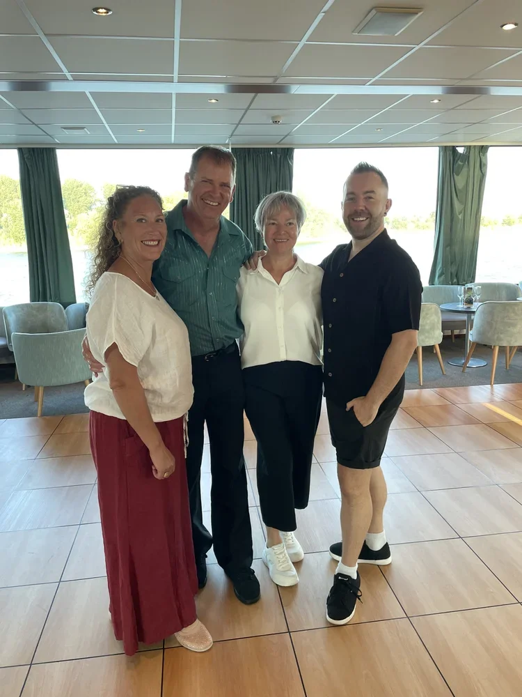 Vier Menschen stehen nah beieinander in einem Raum mit großen Fenstern, die einen Wasserblick zeigen. Sie lachen und posieren für die Kamera, die Gruppenszene wirkt freundlich und gesellig.