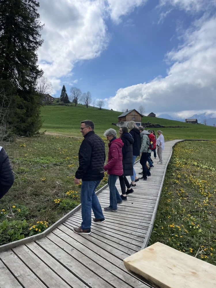 Menschen gehen auf einem hölzernen Steg durch eine grüne Wiese mit gelben Blumen, im Hintergrund sind Häuser und Bäume, bei bewölktem Himmel.