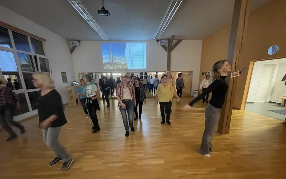 Menschen, die in einem hell erleuchteten Raum tanzen oder spazieren, mit Holzboden, großen Fenstern und einer Projektionswand an der Wand.