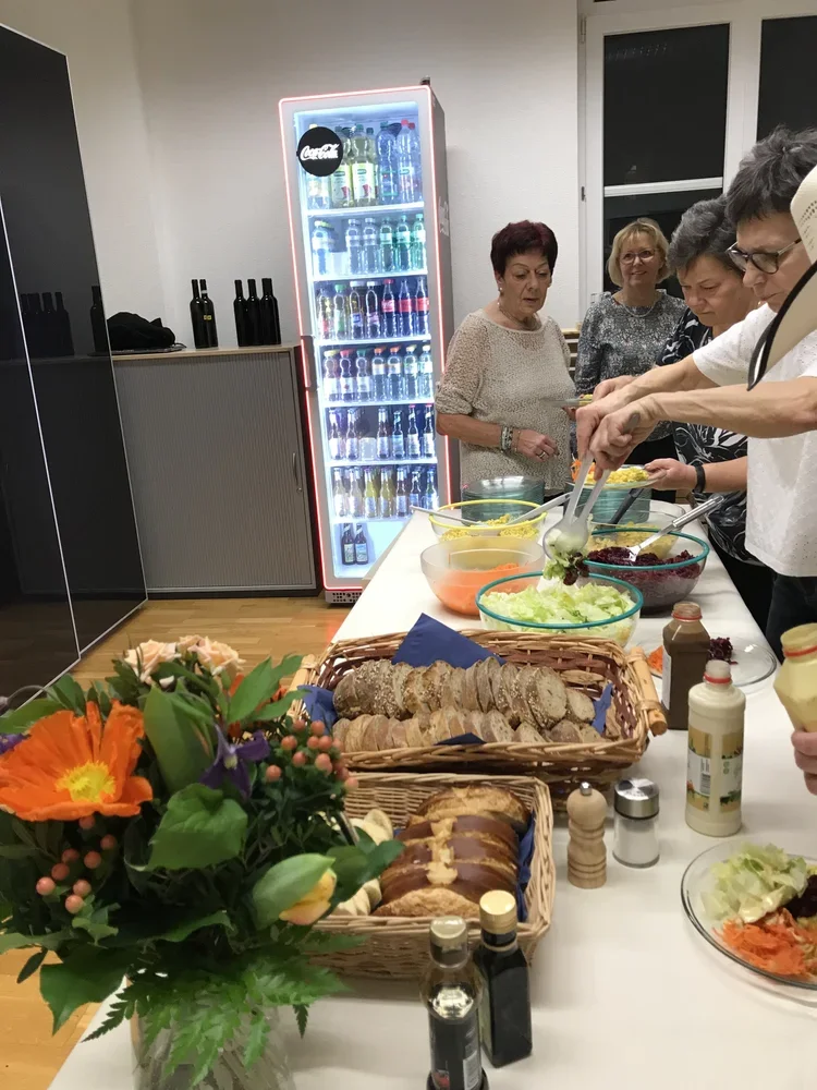 Menschen beim Buffet, mit Salaten, Brot und Getränken, in einem Raum mit Kühlschrank.