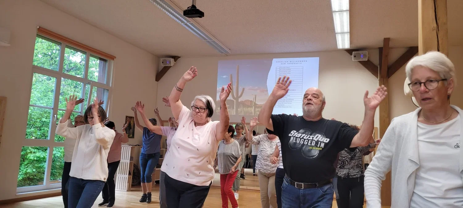 Ältere Menschen machen einen Tanz- oder Bewegungs klasse in einem Gemeinschaftsraum, mit ihren Armen in die Luft und fröhlichen Gesichtsausdrücken, im Hintergrund eine Projektionswand und große Fenster mit grünen Bäumen draußen.