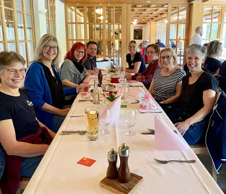 Eine Gruppe von neun Menschen sitzt an einem langen Esstisch in einem Restaurant, alle lachen und haben eine gute Zeit.