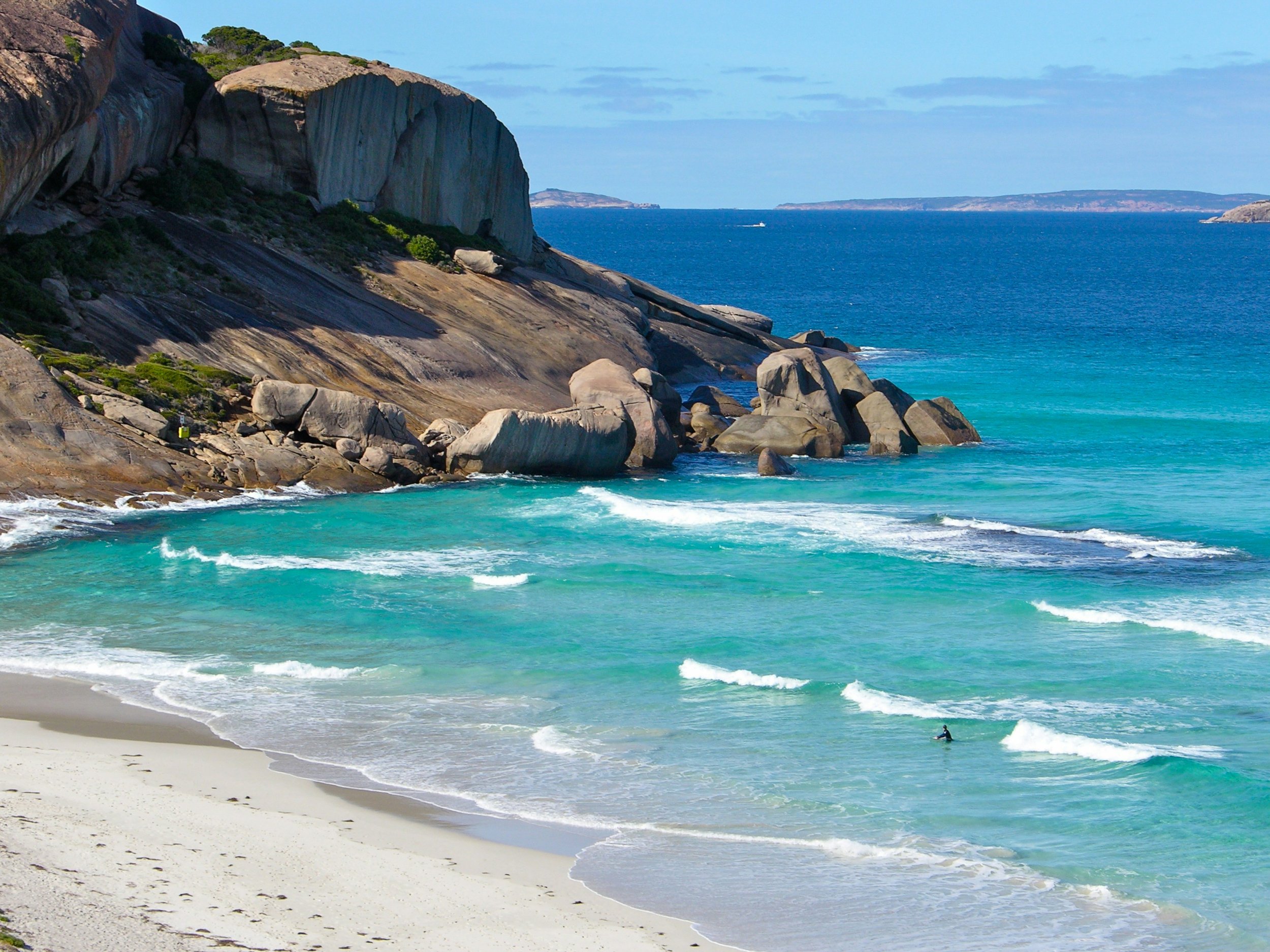 View of a rocky coastline with large granite formations, clear blue ocean waves, and a small surfer in the water near the sandy beach.