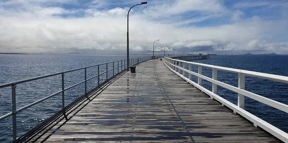 A wooden pier extends over the ocean with white railings on one side and gray railings on the other. There are streetlights along the pier, and it appears to be a cloudy day with some blue sky visible.